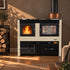 A freestanding wood-burning stove with two glass doors, one showing a visible flame, set in a styled room with wood floors, a log rack, and a copper pot on top. The stove has a cream and black finish with the Hydro brand visible on the front.