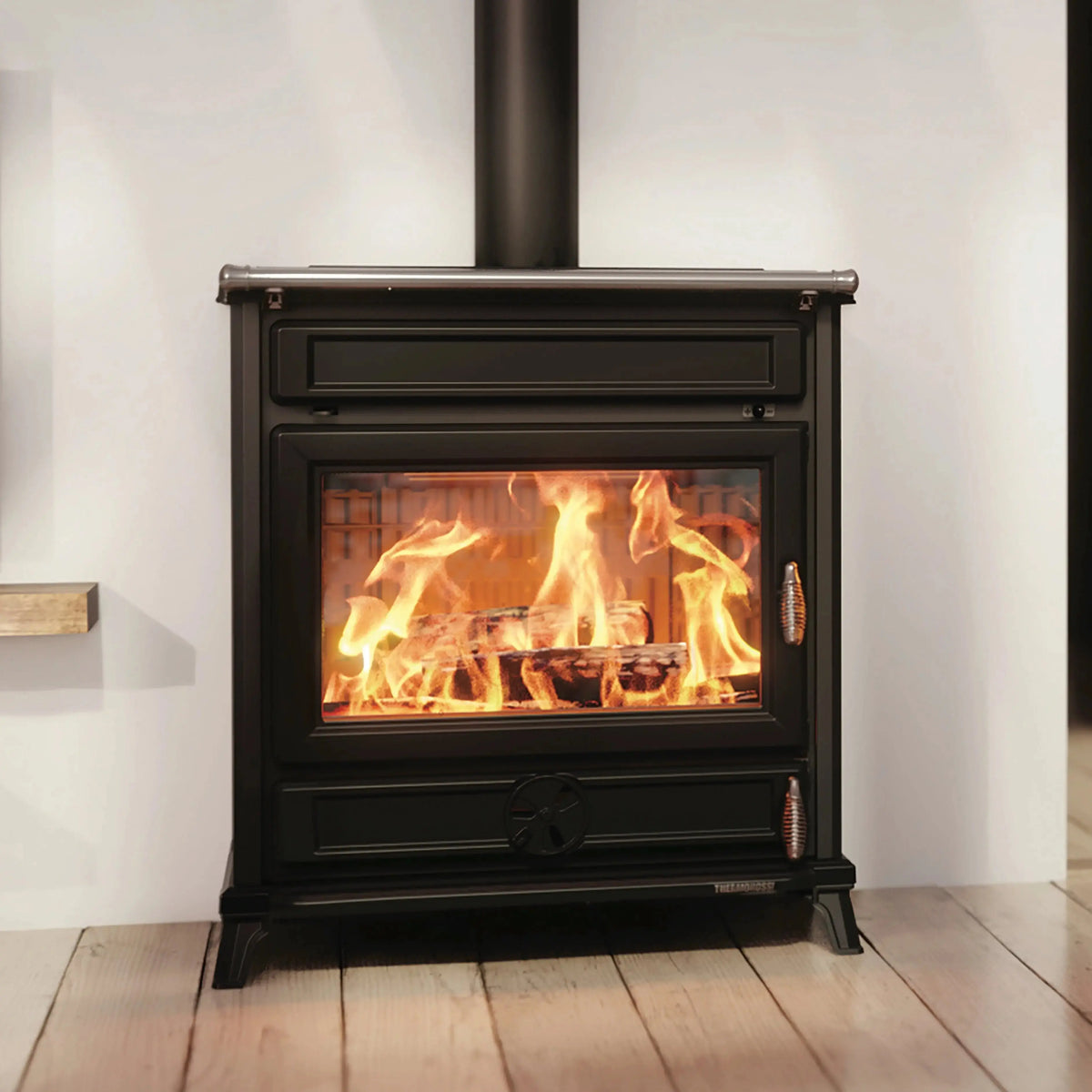 A black wood-burning stove with a visible fire inside, set against a white wall and light wood floor.