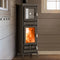 A tall black wood-burning stove with a glass door showing a bright orange fire, set against a brown paneled wall in an indoor room.