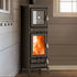 A tall black wood-burning stove with a glass door showing a bright orange fire, set against a brown paneled wall in an indoor room.