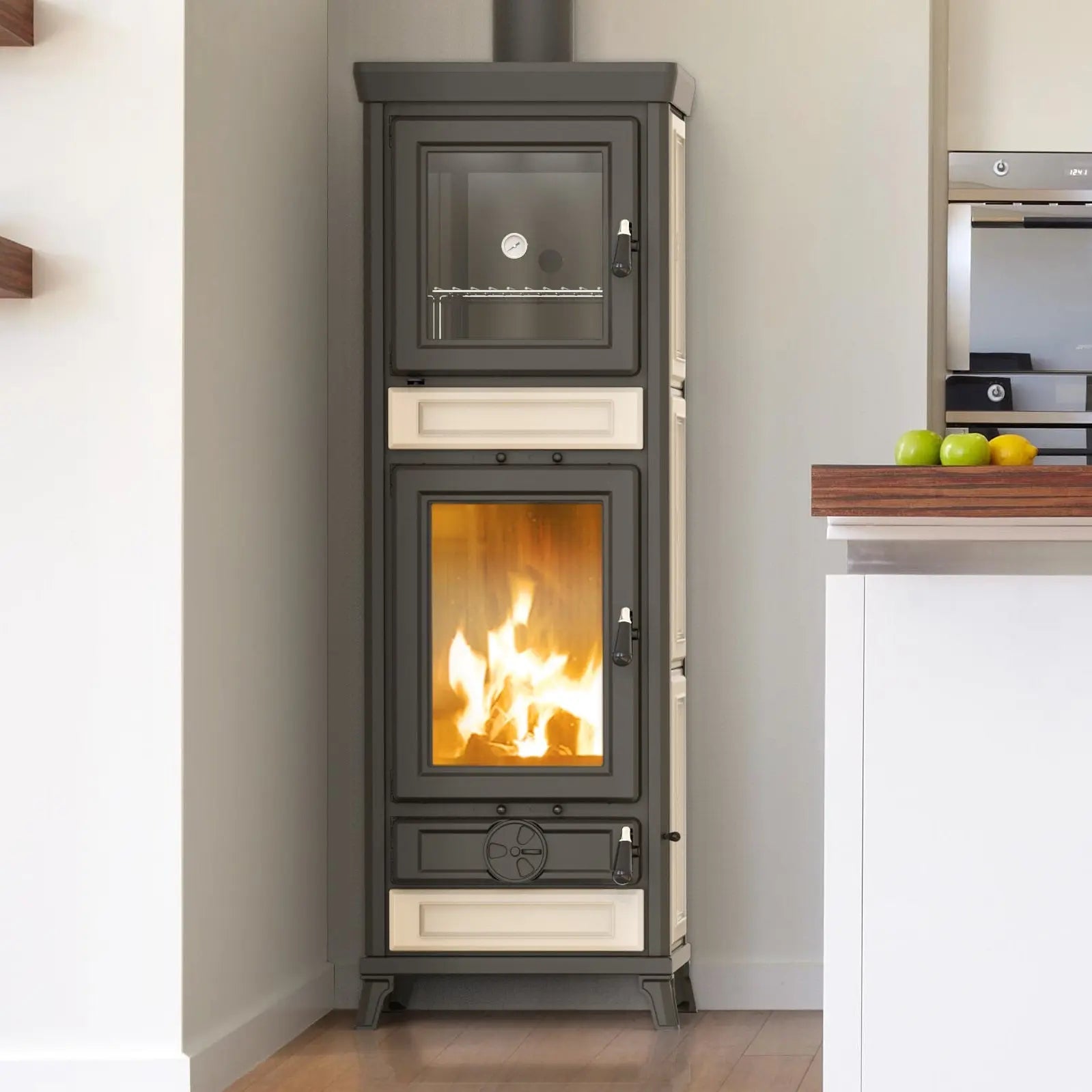A tall freestanding wood-burning stove with a glass door showing a fire inside, set in a real indoor room with a wall, floor, and kitchen area visible.