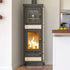 A tall freestanding wood-burning stove with a glass door showing a fire inside, set in a real indoor room with a wall, floor, and kitchen area visible.