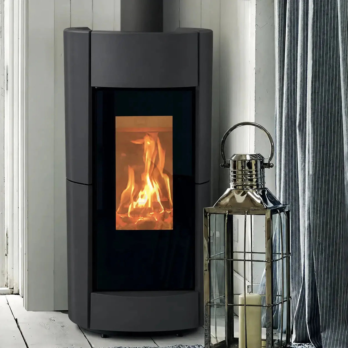 A modern matt black wood-burning stove with a black glass front door showing a glowing orange fire, set against a white paneled wall next to a silver metal lantern.