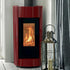 A modern burgundy wood-burning stove with a black glass front showing a glowing fire, set against a white paneled wall next to a silver lantern and striped curtain.