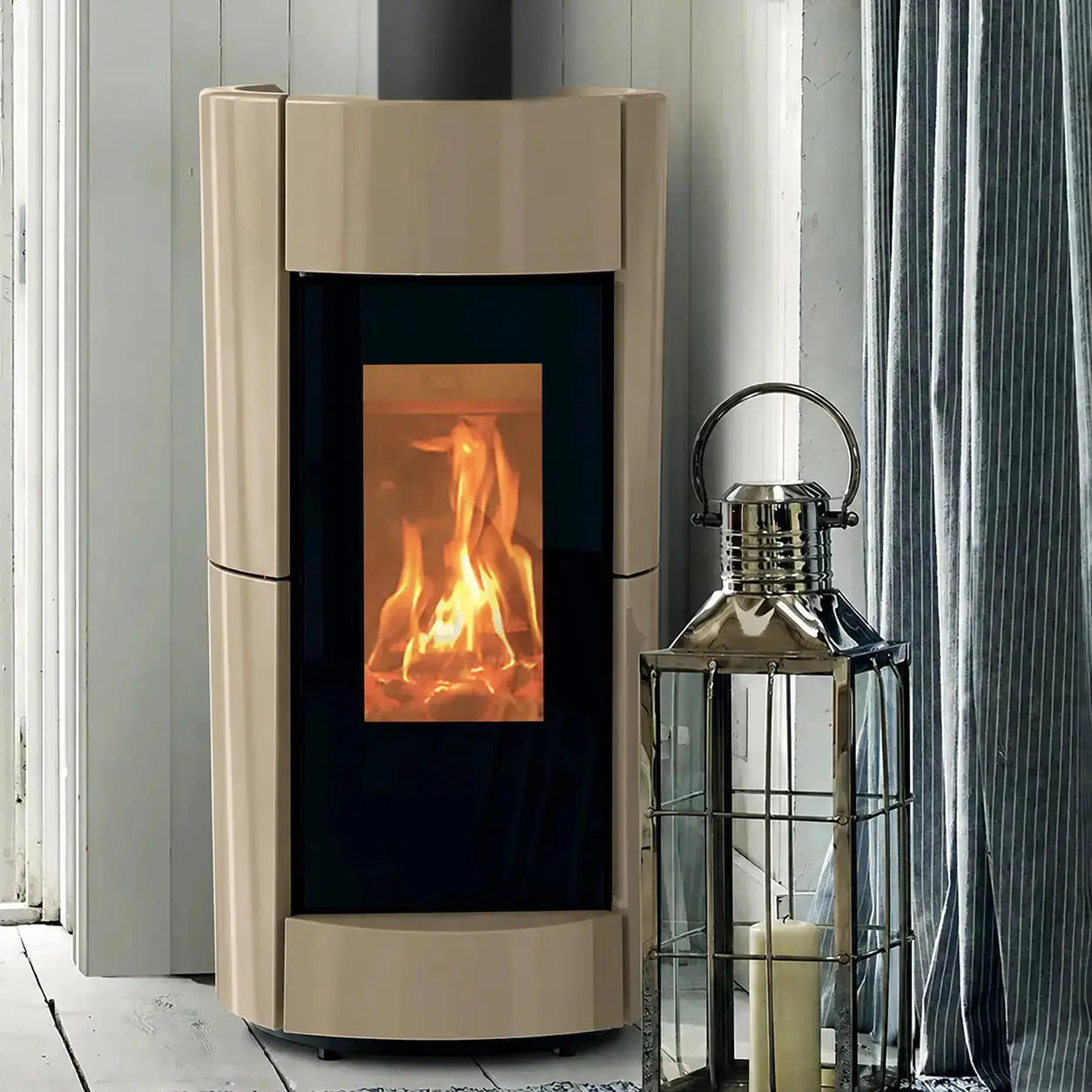 A modern wood-burning stove with a down frame and a black glass front showing a glowing fire, set against a white paneled wall next to a silver lantern and gray curtains.