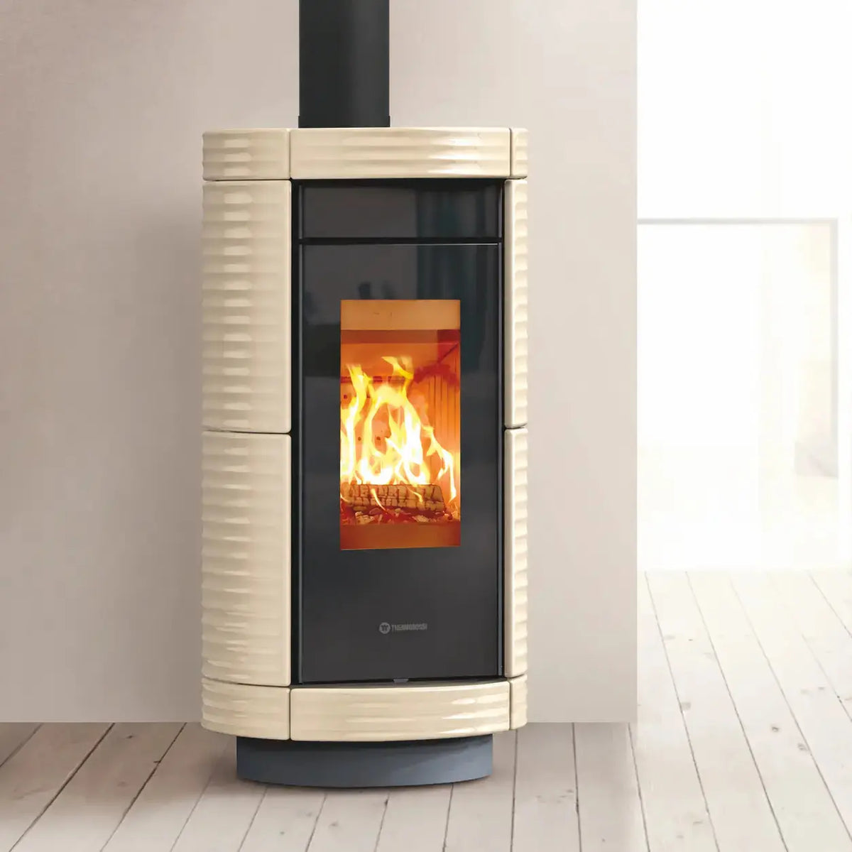 A modern cylindrical wood-burning stove with a cream-colored ribbed ceramic body, black glass door, and a bright orange fire burning inside, set against a minimalist beige wall and light wood floor.