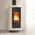 A modern cylindrical wood-burning stove with a cream-colored ribbed ceramic body, black glass door, and a bright orange fire burning inside, set against a minimalist beige wall and light wood floor.