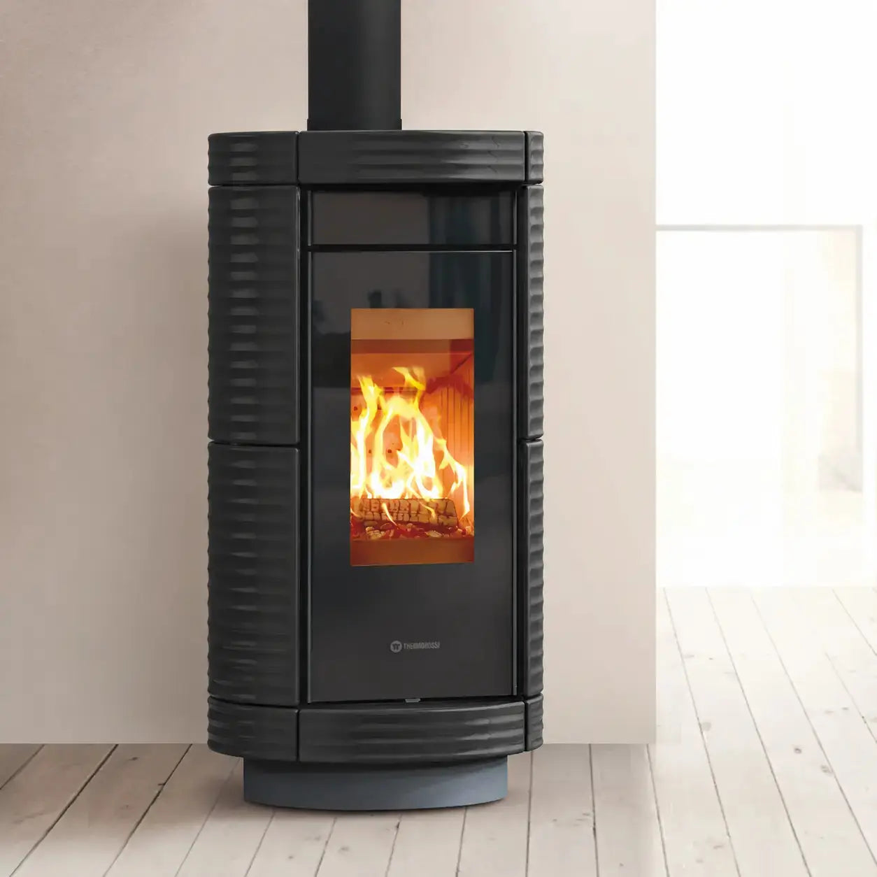 A modern black wood-burning stove with a ribbed ceramic exterior and a glass front showing a warm fire, set against a minimalist beige wall and light wood floor.