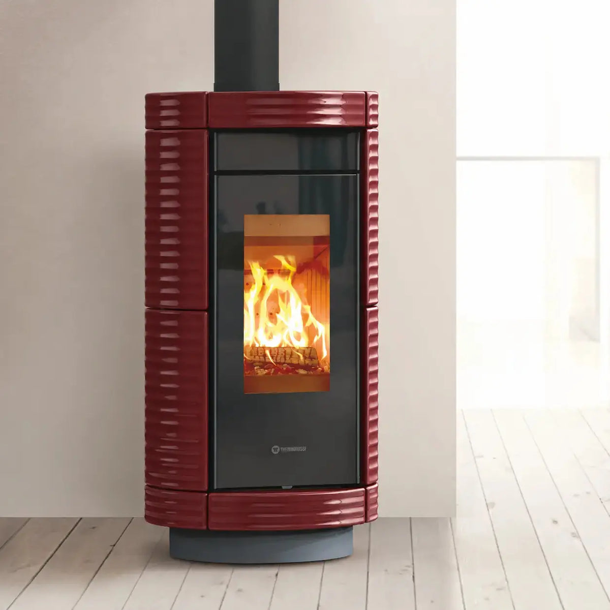 A modern burgundy and black wood-burning stove with a visible fire inside, set against a minimalist beige wall and light wood floor.