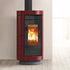 A modern burgundy and black wood-burning stove with a visible fire inside, set against a minimalist beige wall and light wood floor.