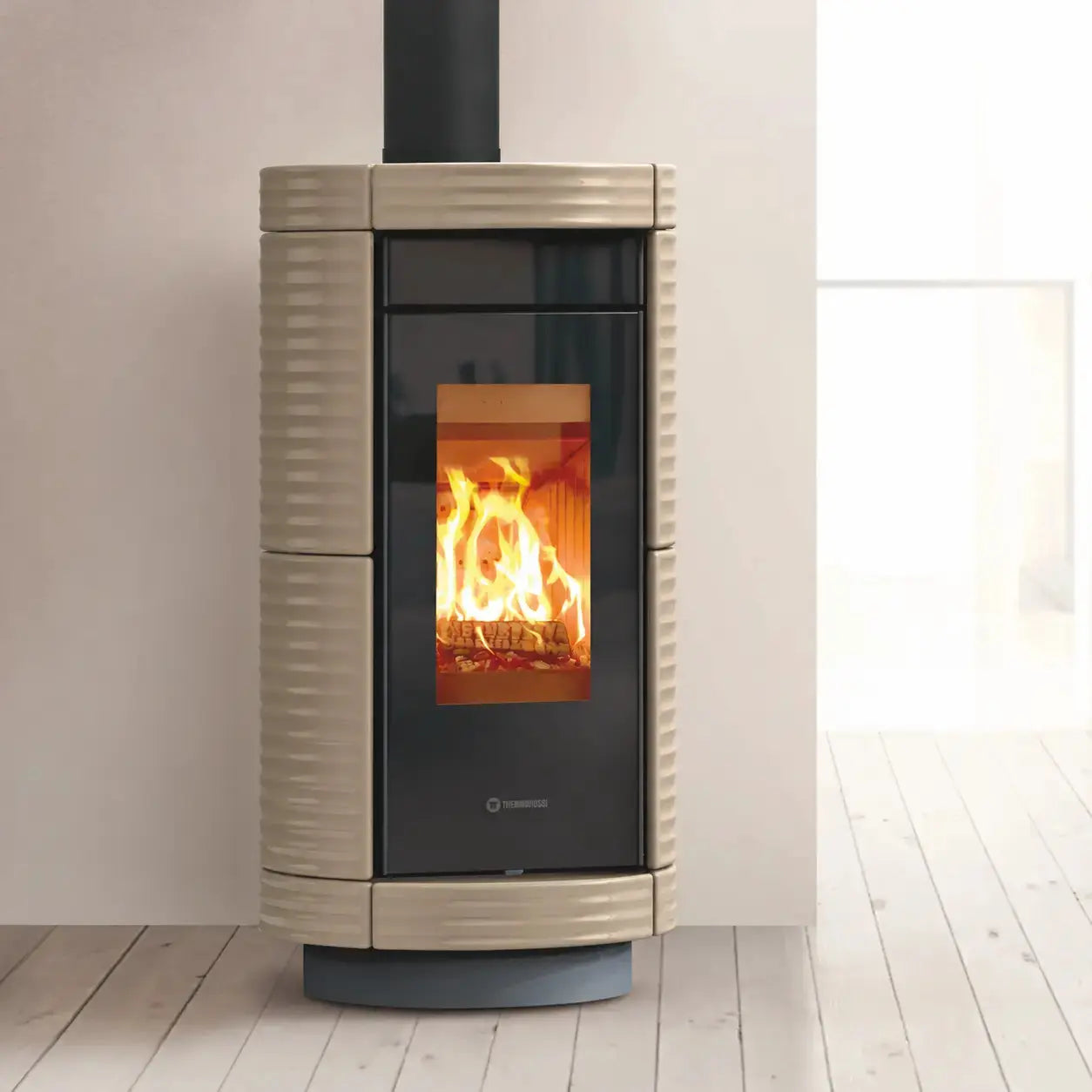 A modern wood-burning stove with a ribbed beige exterior, black glass door, and visible orange fire inside, standing on a light wood floor against a neutral wall.