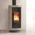 A modern wood-burning stove with a ribbed beige exterior, black glass door, and visible orange fire inside, standing on a light wood floor against a neutral wall.
