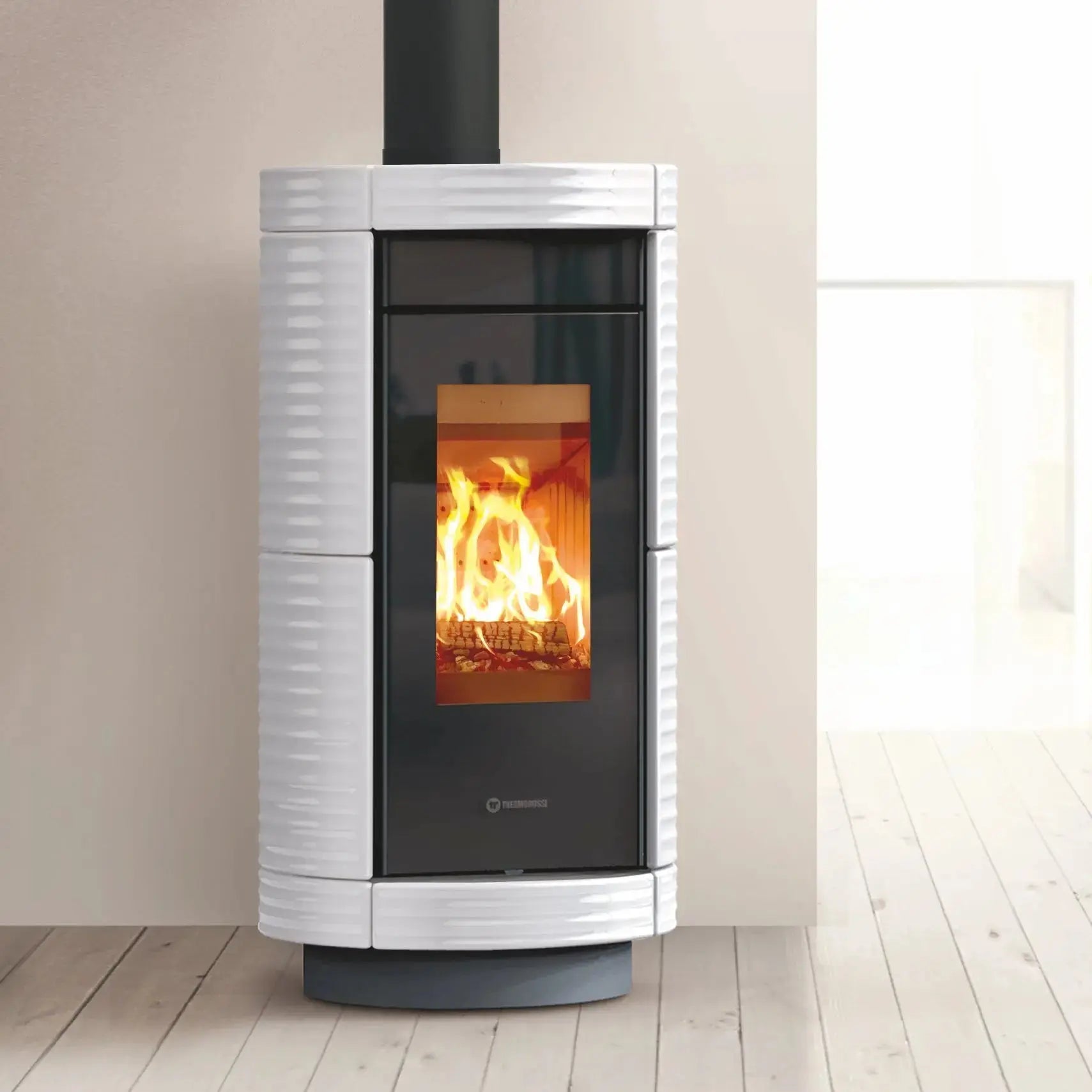 A modern wood-burning stove with a white ribbed ceramic body, black glass door, and visible orange fire, set against a neutral beige wall and light wood floor.