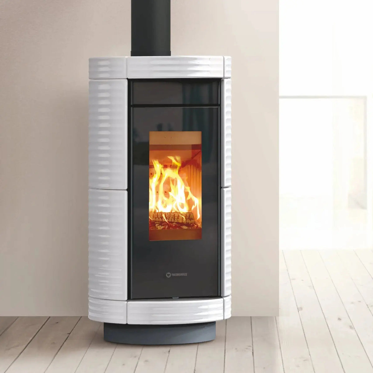 A modern wood-burning stove with a white ribbed ceramic body, black glass door, and visible orange fire, set against a neutral beige wall and light wood floor.