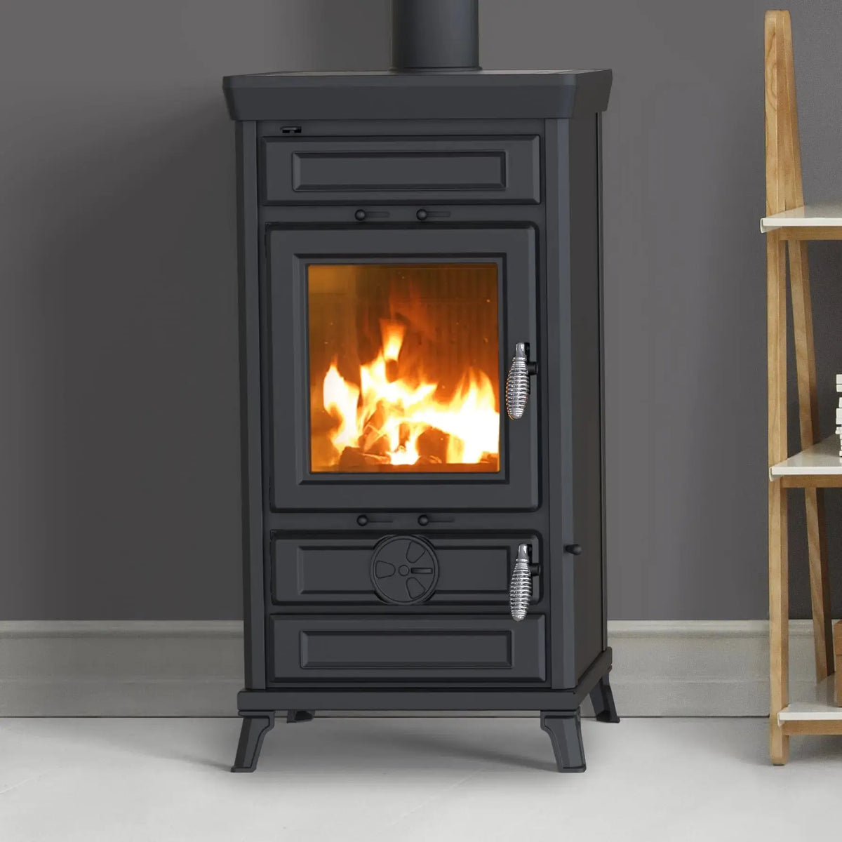 A black metal wood-burning stove with a visible orange fire inside, set against a dark gray wall and light floor. A small wooden shelf is partially visible on the right side.