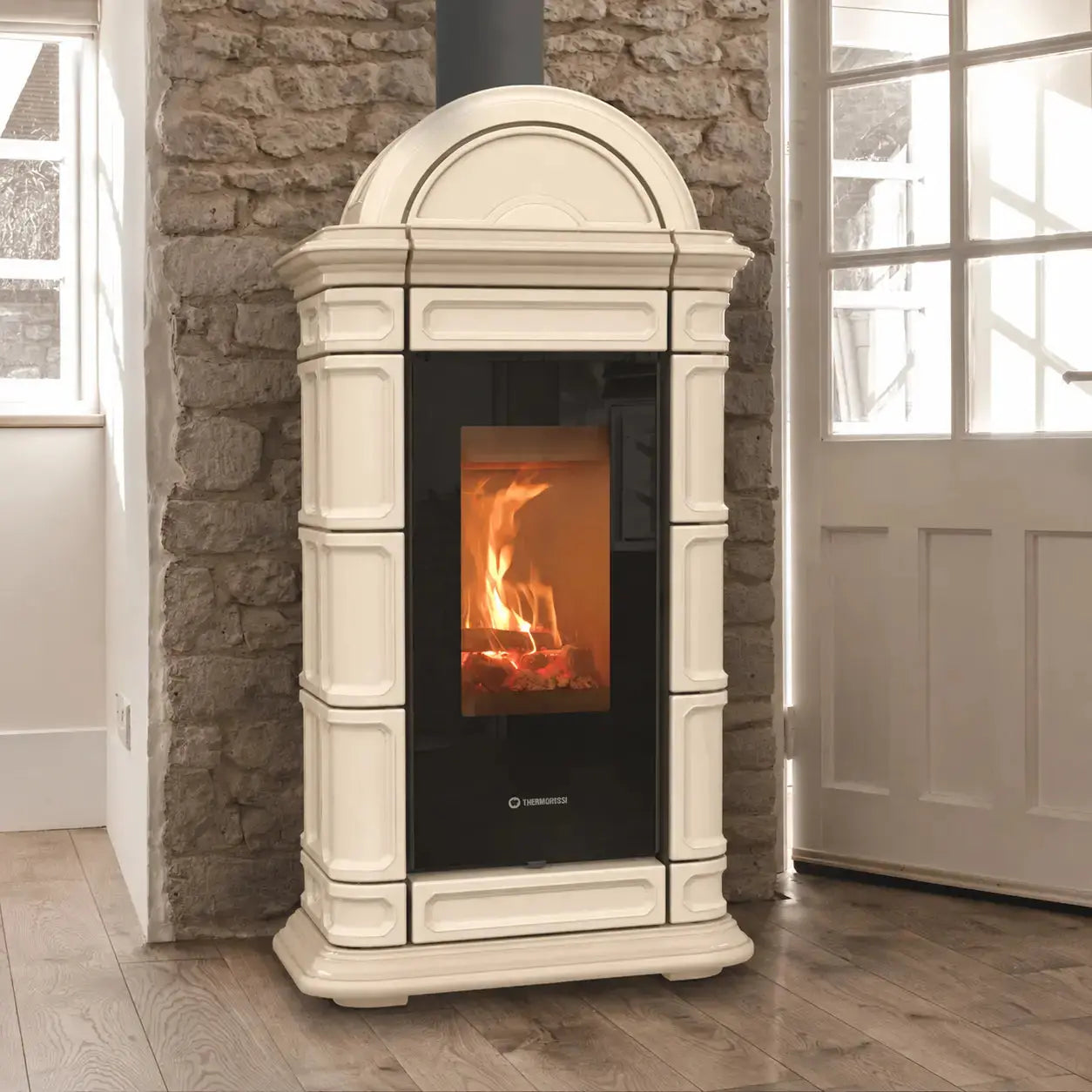 An beige freestanding wood-burning stove with a black glass front door showing a fire, set against a rustic stone wall and wood floor.