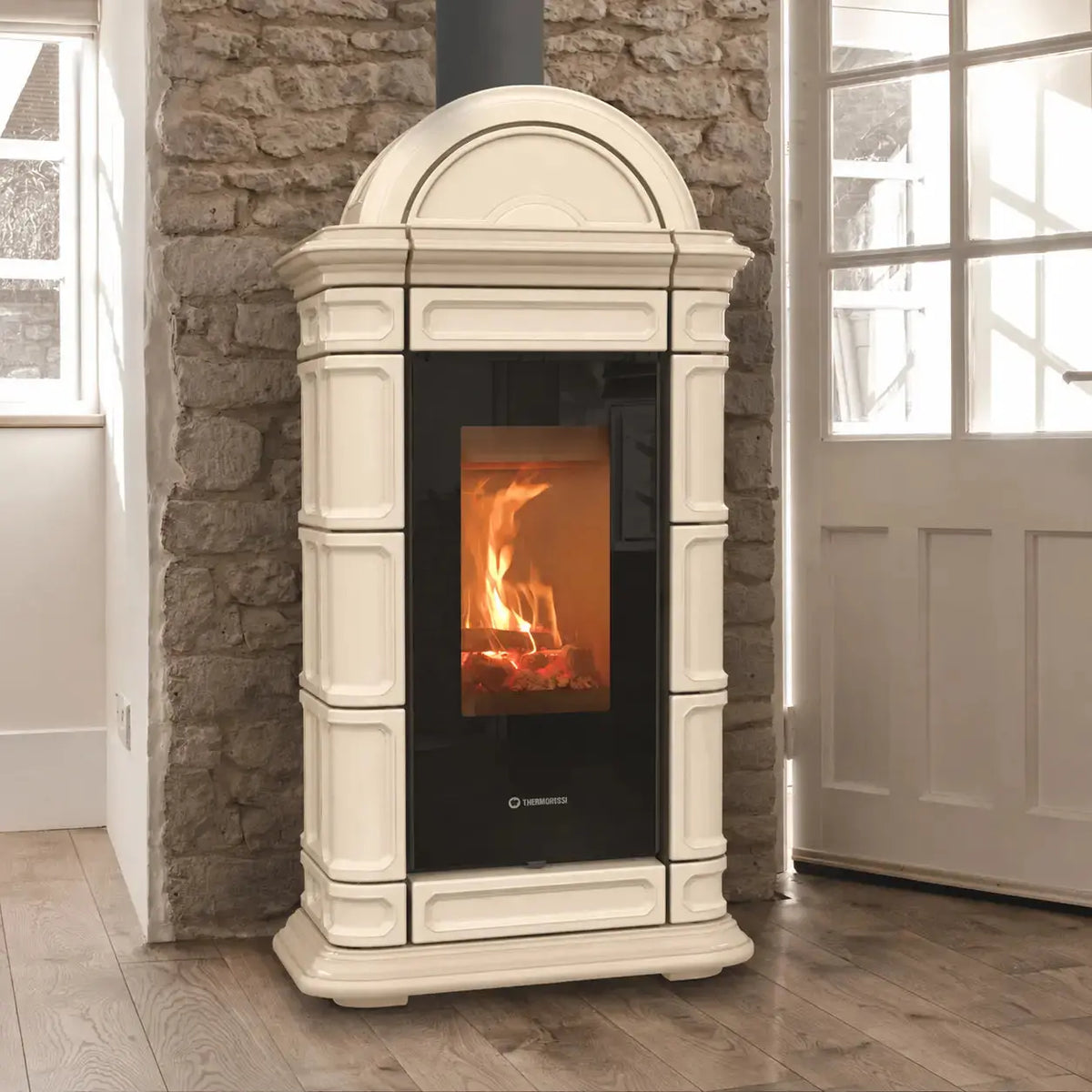 An beige freestanding wood-burning stove with a black glass front door showing a fire, set against a rustic stone wall and wood floor.