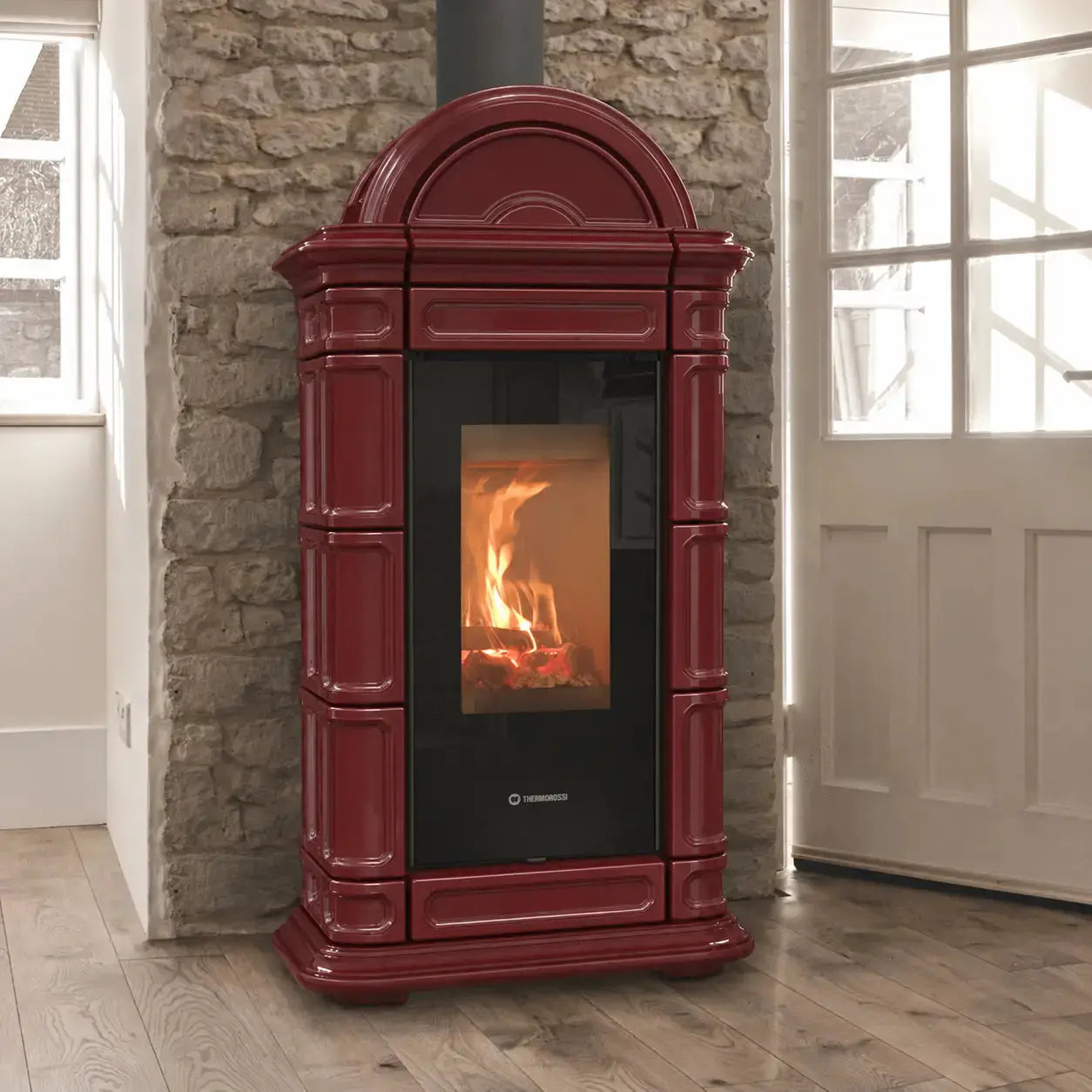 A burgundy freestanding wood-burning stove with a black glass door showing a fire, set against a stone wall in a room with wooden floors and a white door.
