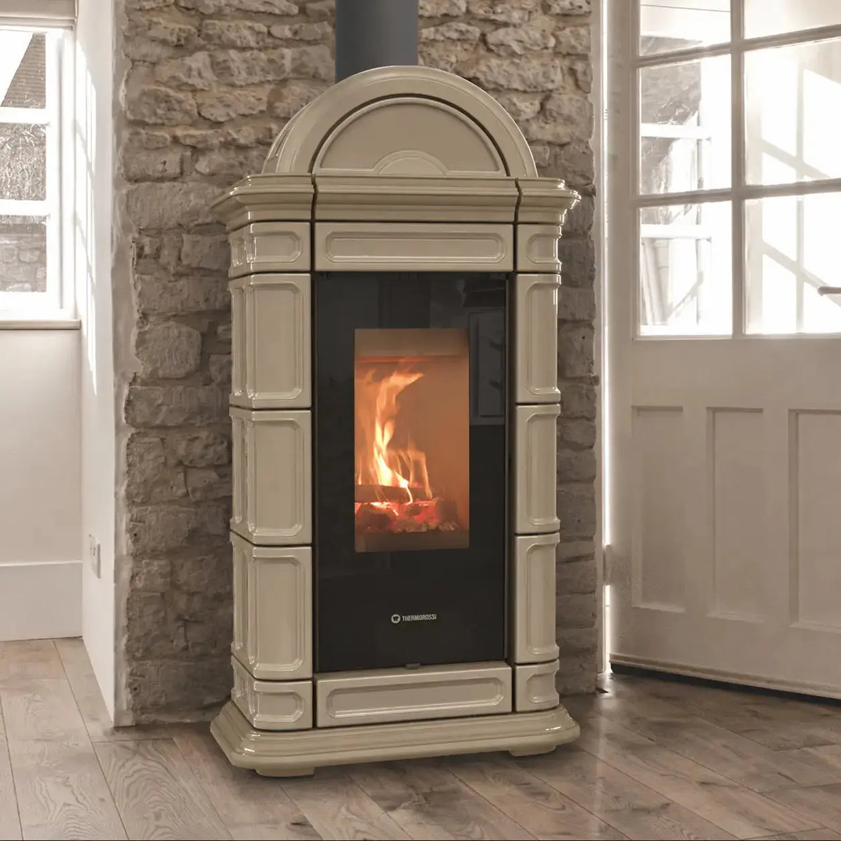 A freestanding wood-burning stove with a down colored ornate frame and a black glass front door showing a glowing fire, set against a stone wall in a room with wood floors and a white door.
