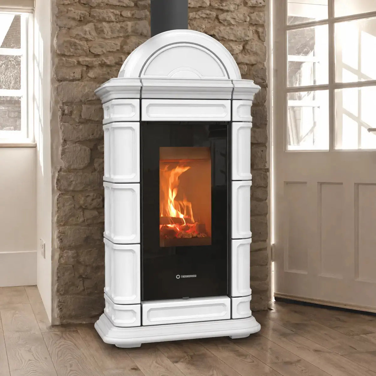 A white freestanding wood-burning stove with a black glass door showing a fire, set against a stone wall and wooden floor.