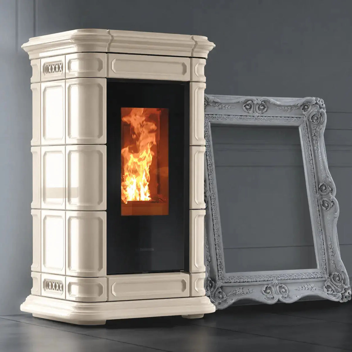 A beige freestanding wood-burning stove with a black glass door showing a glowing fire, photographed indoors against a dark gray wall. A large ornate white picture frame leans against the stove as a prop.