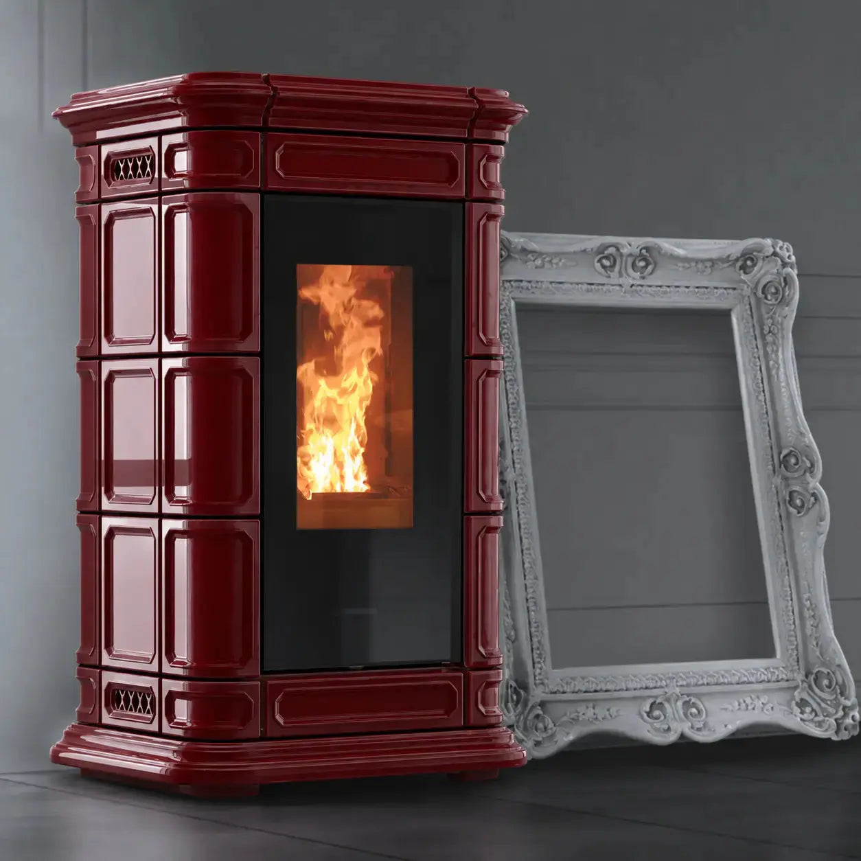 A burgundy freestanding wood-burning stove with a black glass door showing a glowing fire, photographed indoors against a dark gray wall. A large ornate white picture frame leans against the stove as a prop.