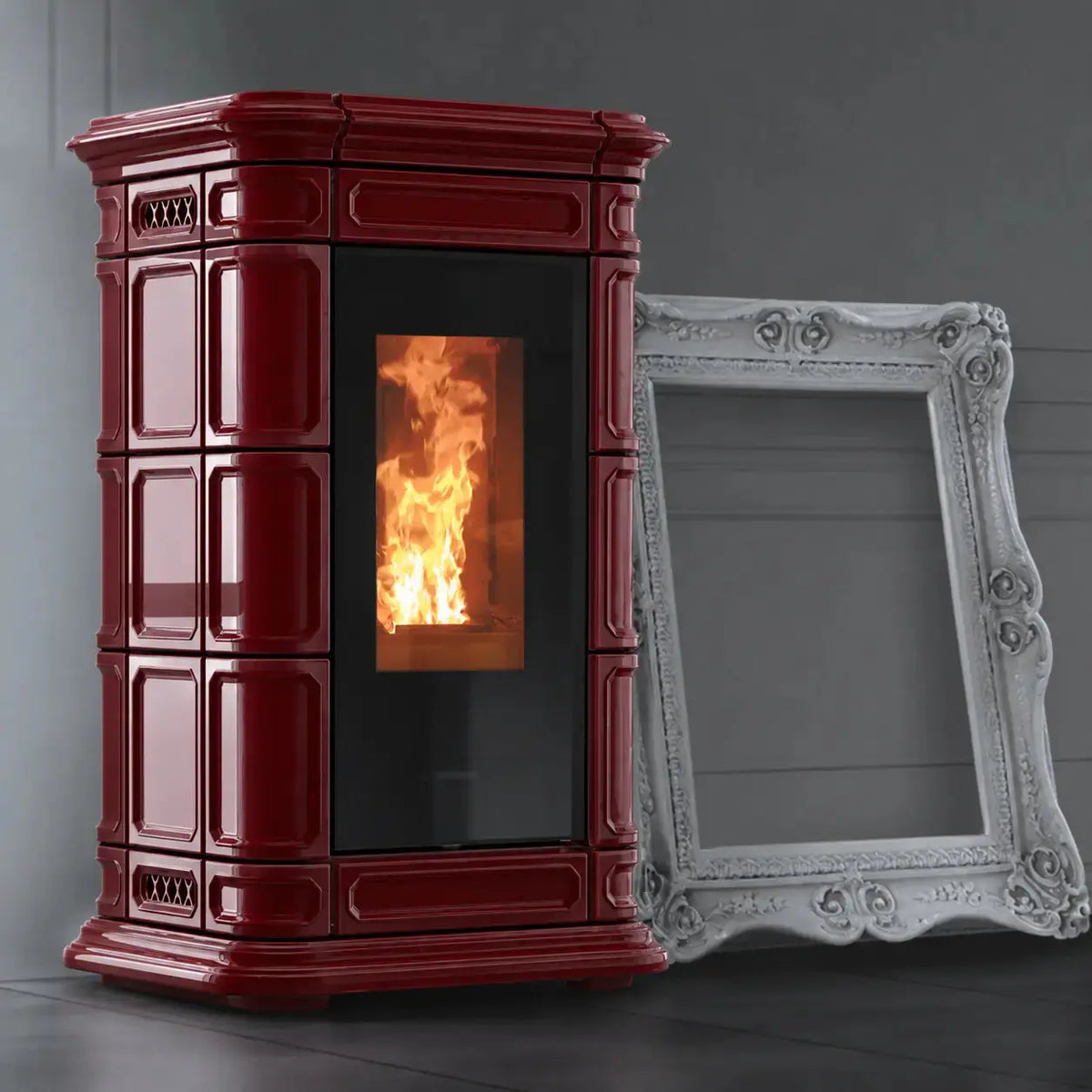 A burgundy freestanding wood-burning stove with a black glass door showing a glowing fire, photographed indoors against a dark gray wall. A large ornate white picture frame leans against the stove as a prop.