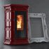 A burgundy freestanding wood-burning stove with a black glass door showing a glowing fire, photographed indoors against a dark gray wall. A large ornate white picture frame leans against the stove as a prop.