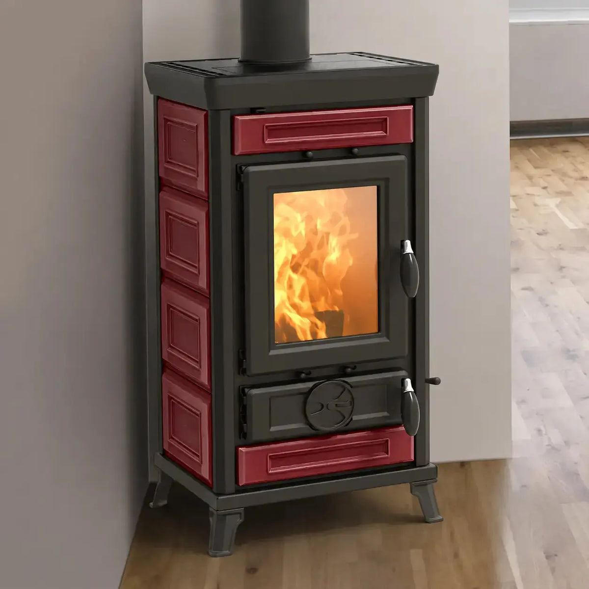 A freestanding wood-burning stove with a black cast iron body and red tile fronts, shown standing on a wood floor against a light wall with a fire visible through the glass door.