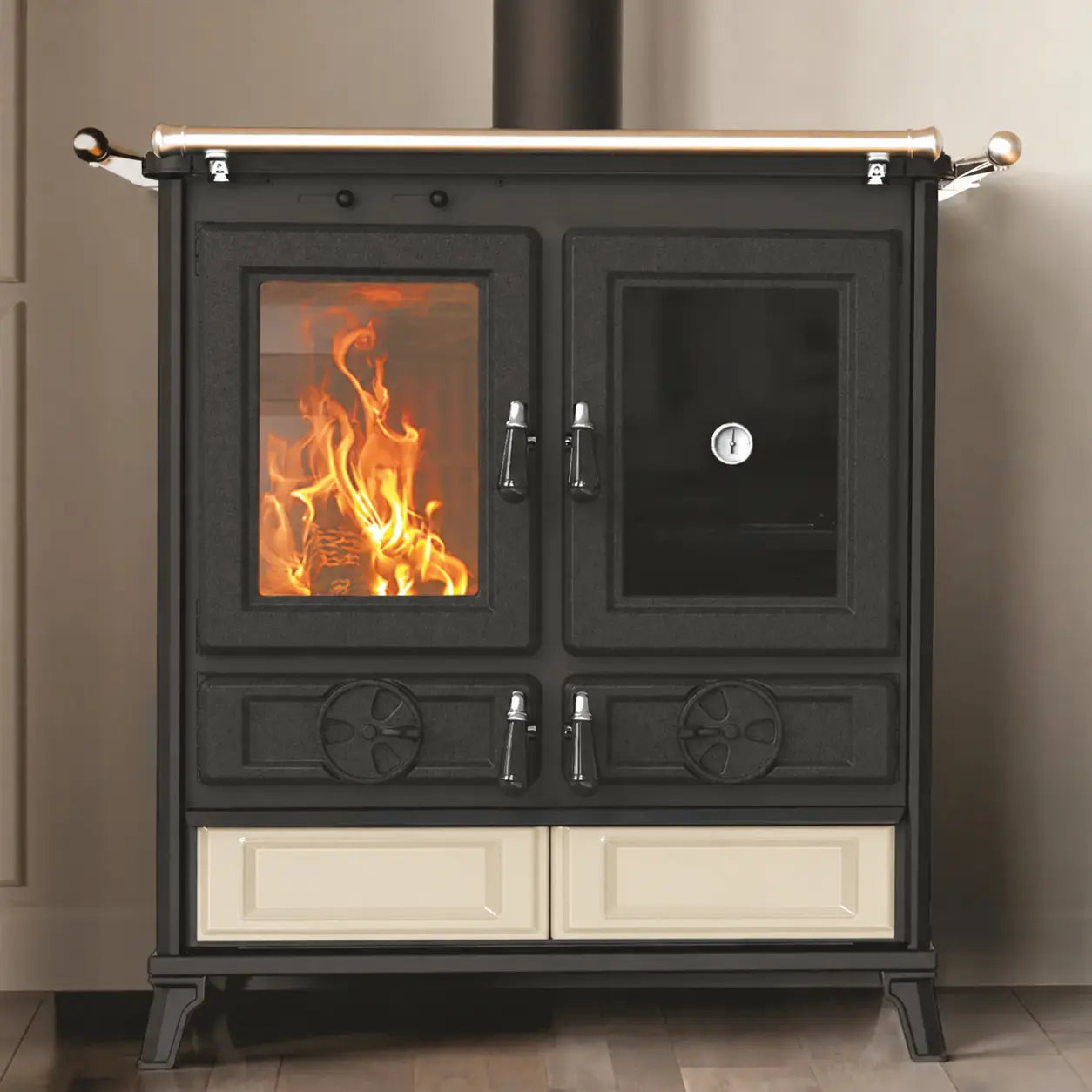 A black cast iron wood-burning stove with a glass front door showing a fire, a side door with a glass window, cream-colored lower drawers, and a brass top bar, set against a neutral beige wall and wooden floor.